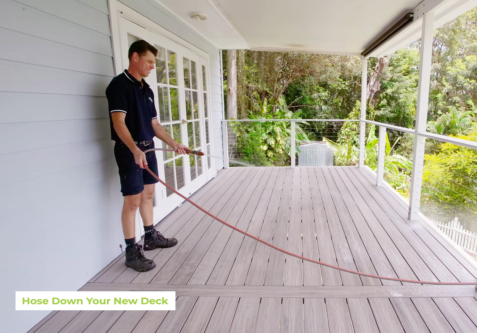 Man rinsing a light grey composite deck with a garden hose on a covered balcony surrounded by greenery.