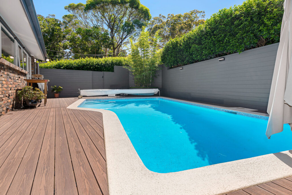 A cozy pool area with composite decking, featuring a clean, rectangular pool surrounded by greenery and tall hedges. The deck leads up to the pool, with a relaxing ambiance created by the surrounding plants and soft sunlight. The pool's clear blue water contrasts with the earthy tones of the wooden deck.