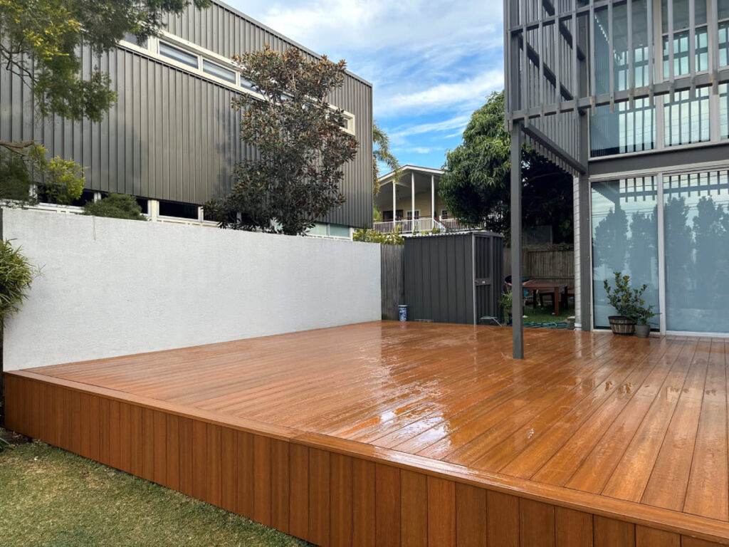 Modern backyard with freshly installed tawny brown composite decking, bordered by a white wall, plants, and contemporary house exterior.