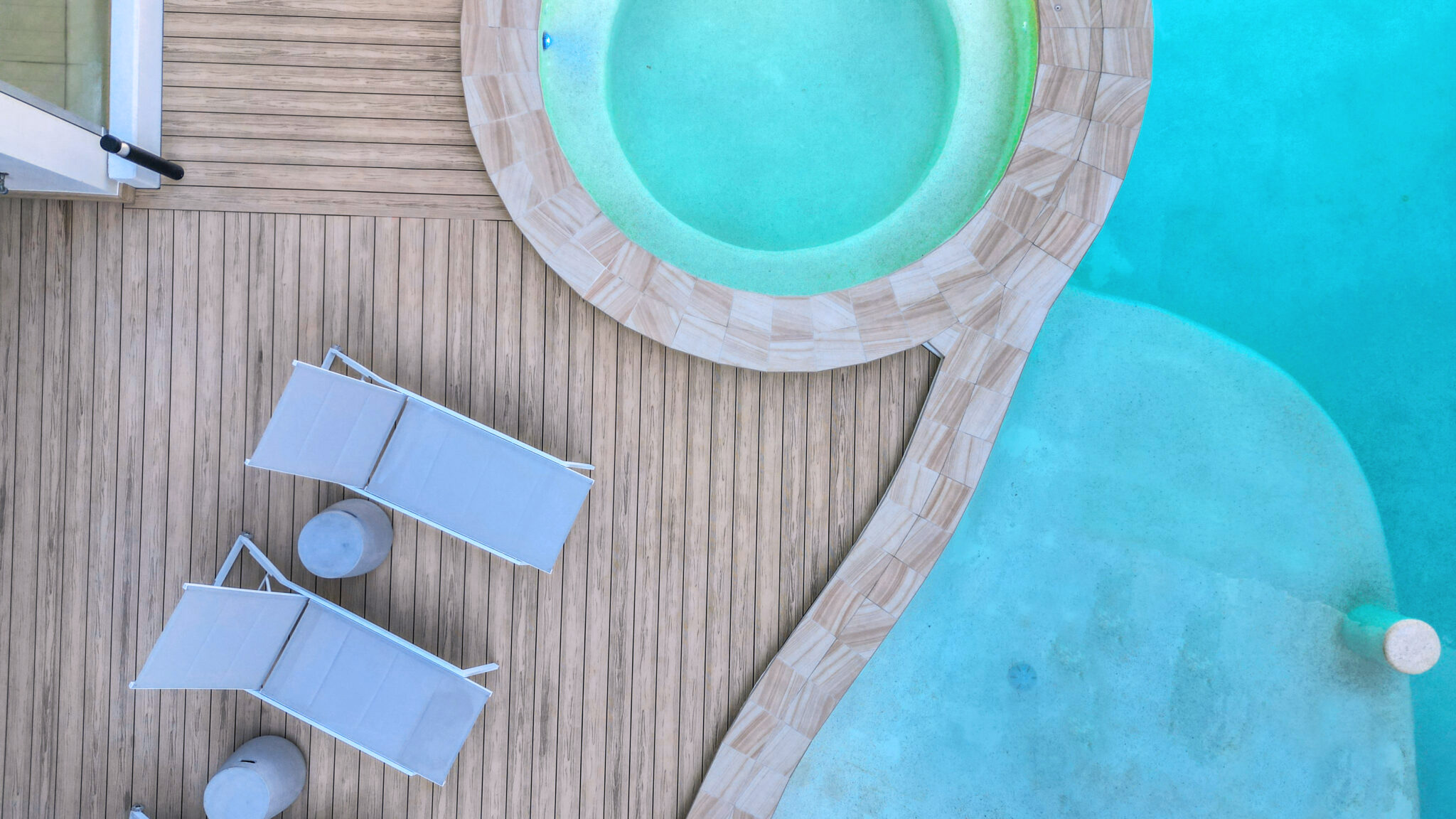 An aerial view of a pool area with composite decking, featuring two white lounge chairs and small round tables. The pool has a circular shape with light-colored tiles around the edge, and the water appears clear and inviting. The wooden deck creates a clean, modern look surrounding the pool area.