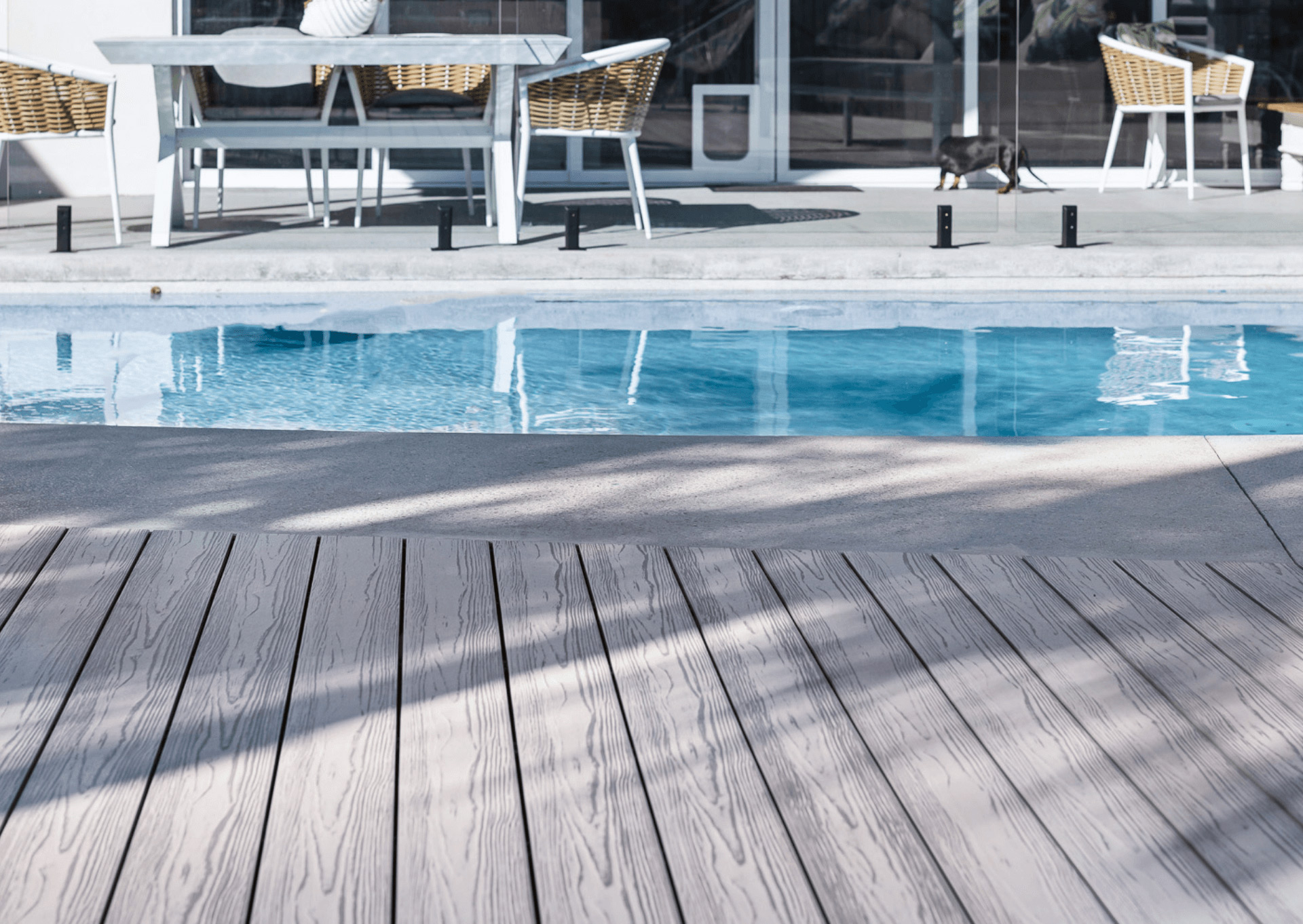 A close-up view of a modern outdoor area featuring light-colored composite decking, leading up to a pool. The deck is smooth and clean, with visible wood grain patterns. In the background, there is outdoor furniture with woven chairs and a table, and a cat is seen walking beside the pool. The pool's clear water reflects the surroundings, creating a serene atmosphere.
