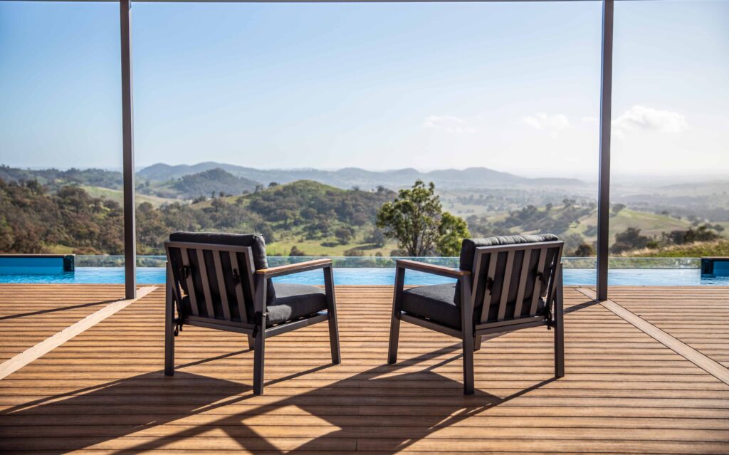 A scenic view of two wooden lounge chairs with dark cushions, placed on a composite deck overlooking a beautiful landscape of rolling hills. The deck is near an infinity pool, and the sunlight casts long shadows on the floor. The clear blue sky enhances the peaceful, serene atmosphere.