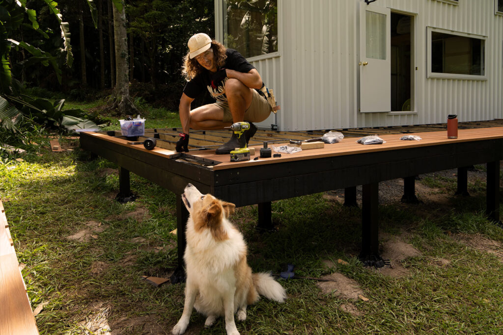 DIY builder working on a composite decking installation beside a tiny house in Byron Bay, with tools spread out and a dog watching from below the raised platform.