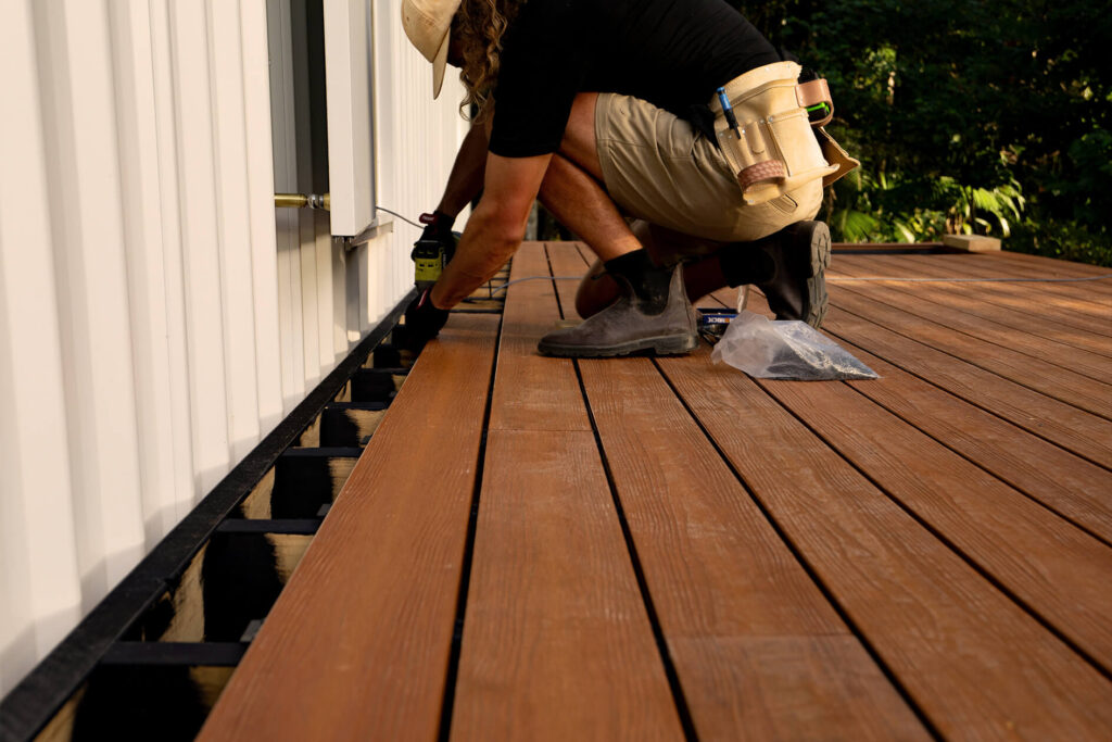 Close-up of a DIY builder fixing the final Titanium Tawny composite decking board against a tiny house wall in Byron Bay, using hidden fasteners for a clean edge finish.