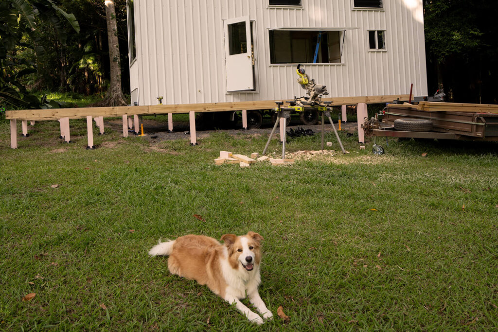DIY composite decking construction in progress for a tiny house in Byron Bay, with tools and timber framing set up and a dog relaxing on the grass nearby