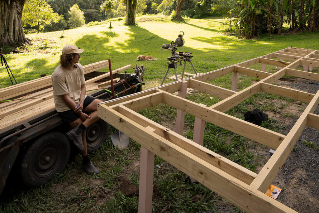DIY composite decking frame construction for a tiny house in Byron Bay, with a builder resting on a trailer and a dog lounging in the sunlit grassy yard.