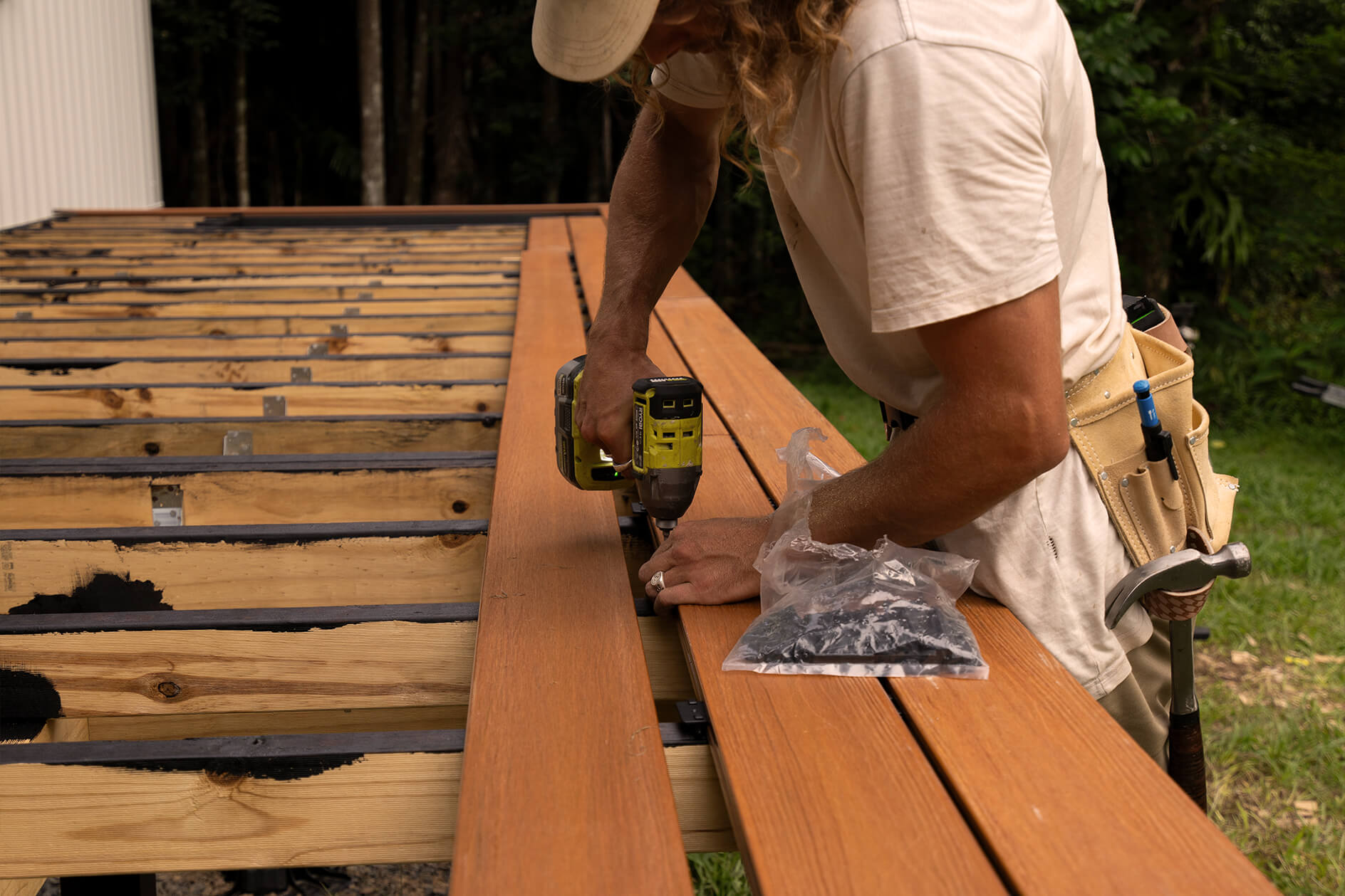 Close-up of DIY builder using a drill to install composite decking boards for a tiny house in Byron Bay, securing Titanium Tawny boards over a timber subframe.