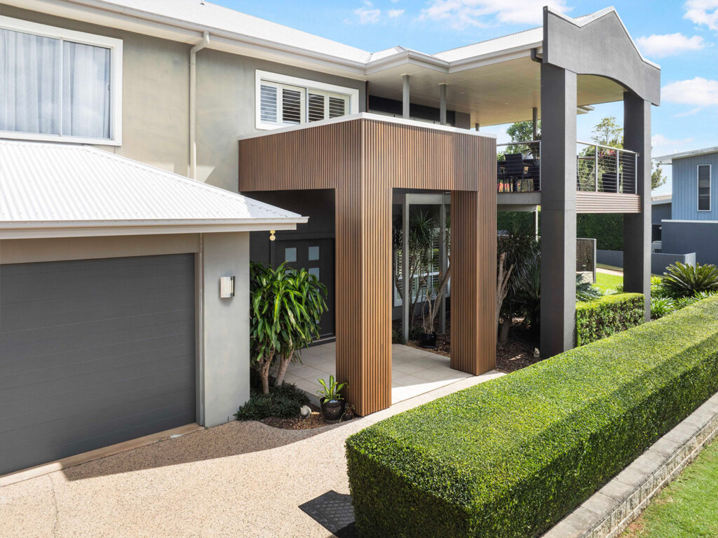 Entryway with Country Teak composite cladding, framed by tiled flooring, lush hedges, and a modern grey garage.