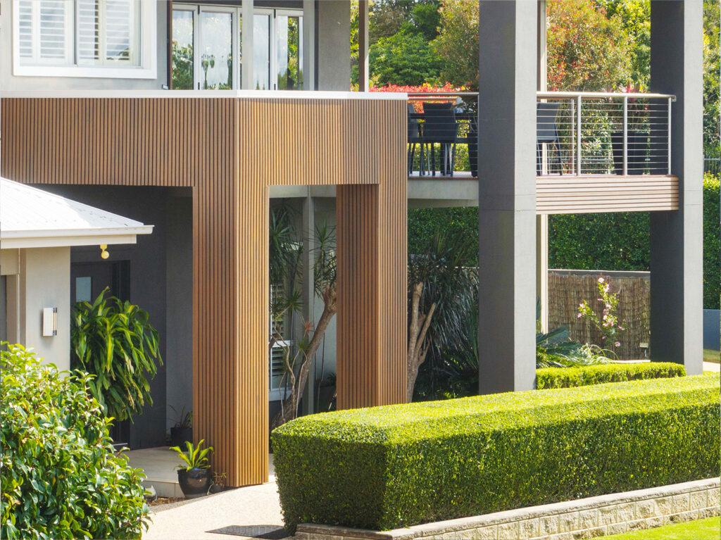 Detailed view of entry framed in Country Teak composite cladding, with vertical lines, potted plants, and vibrant garden hedges