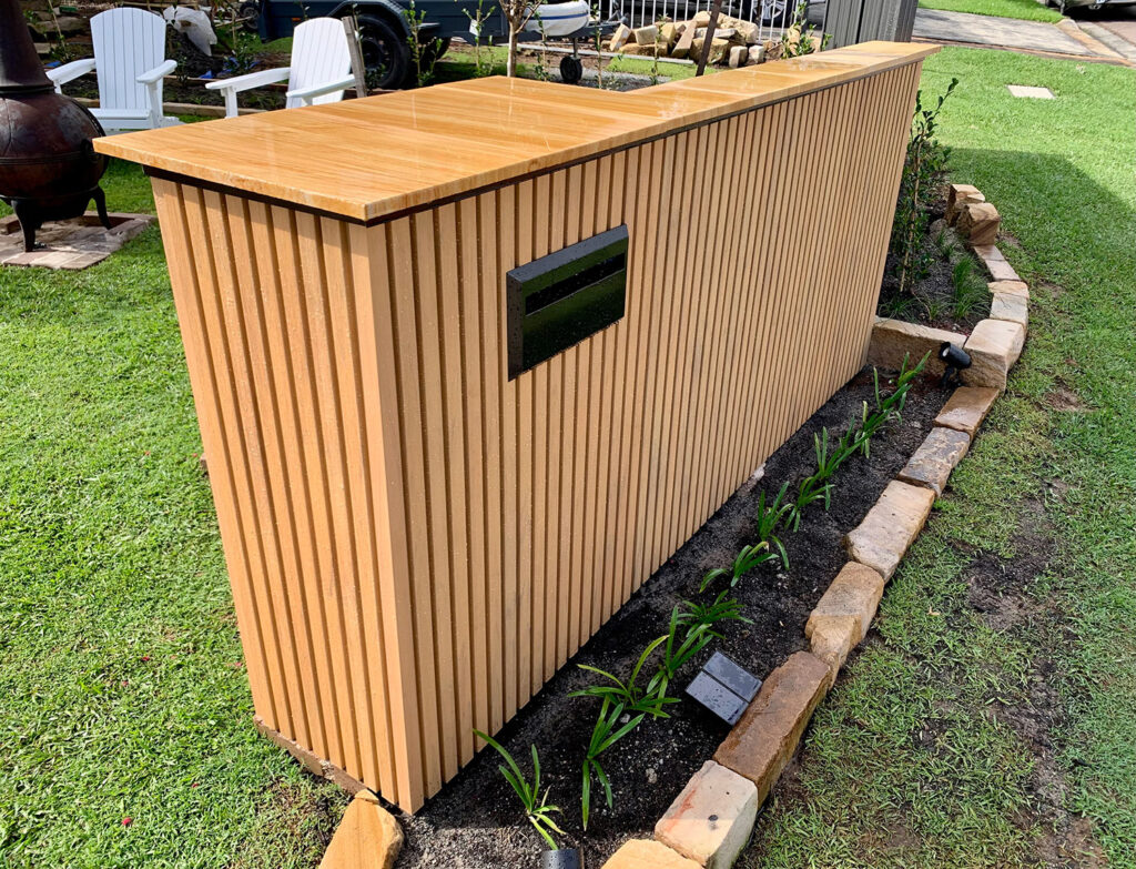 Modern outdoor letterbox clad in vertical slat composite cladding with a timber-look finish, integrated into a landscaped garden with stone borders and new plantings.