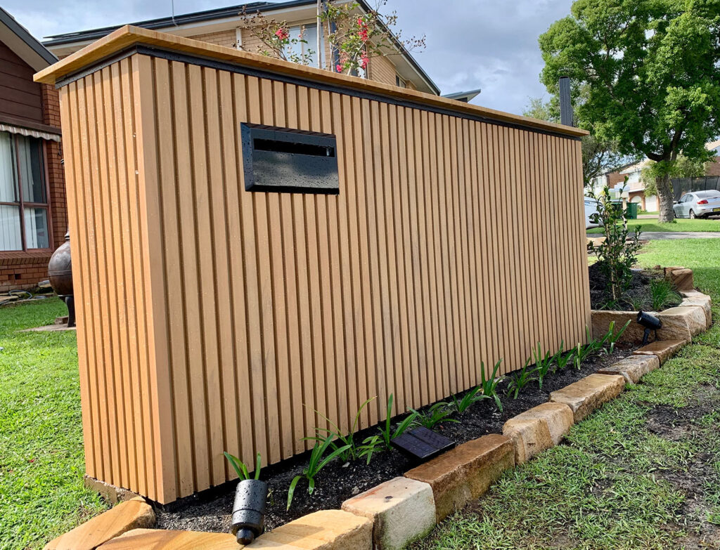 Street-facing view of a modern letterbox structure clad in vertical composite cladding with timber grain finish, black mail slot, and garden edging with young plants and landscape lighting.