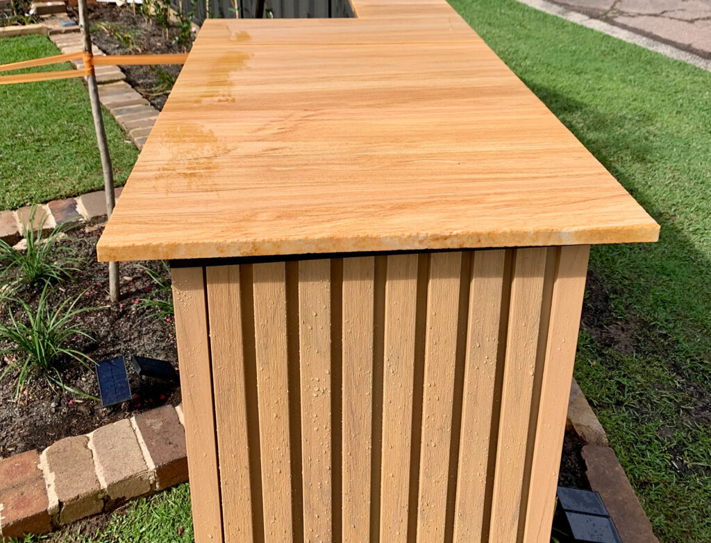 “Close-up of timber-look letterbox finished with vertical groove composite cladding and stone slab top, surrounded by garden edging and solar lights