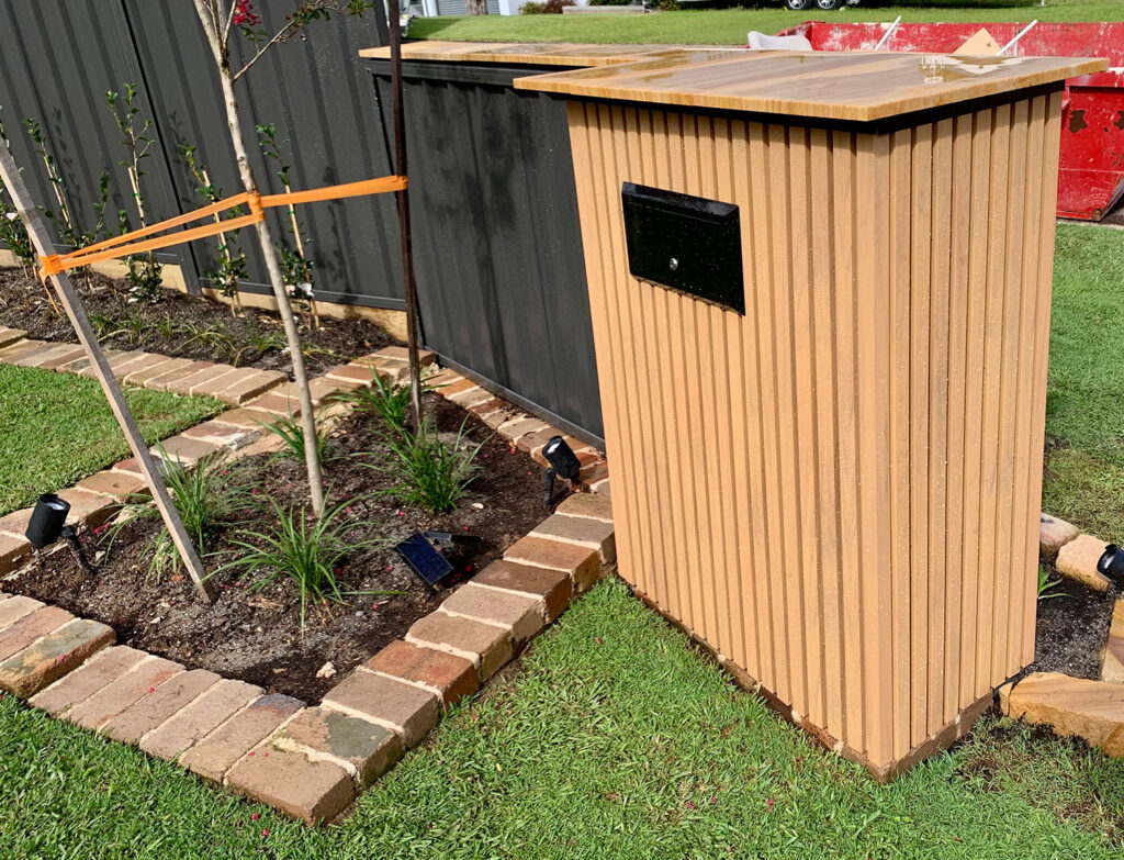 Rear view of a dual-access letterbox structure clad in vertical slat composite cladding, positioned beside a garden bed with young trees, solar lights, and brick edging.