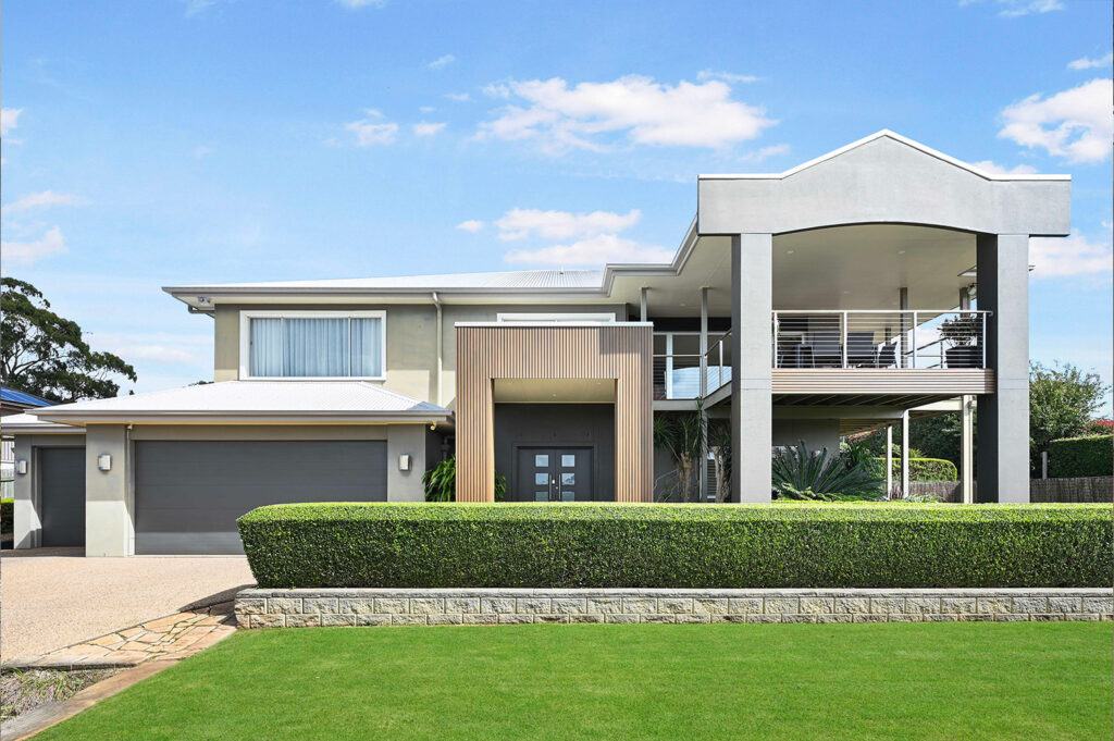 Front elevation of a modern home showcasing Country Teak composite cladding at the entrance, with a balcony, triple garage, and manicured lawn.