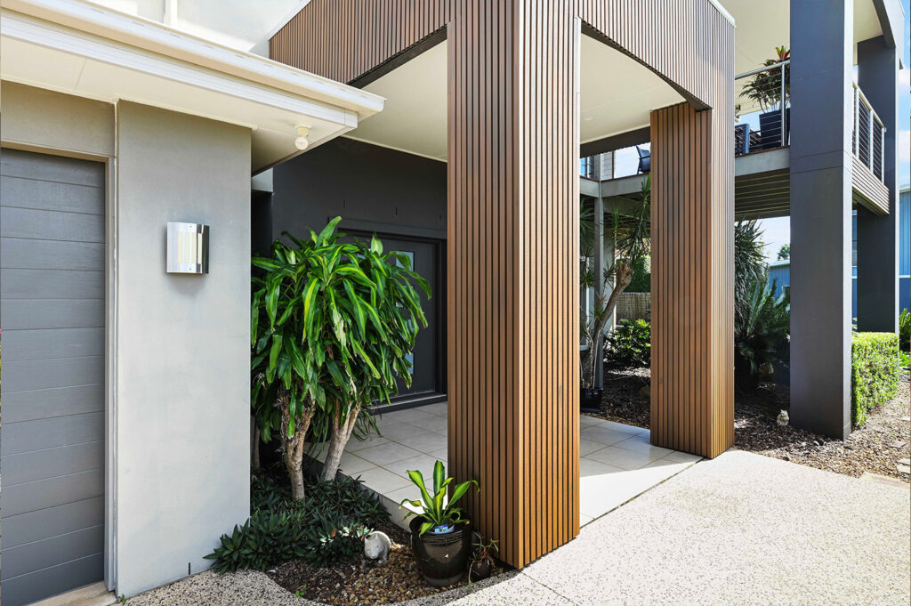 Ground-level view of Country Teak composite cladding entry pillars with tiled floor, potted plants, and a modern dark entry door.