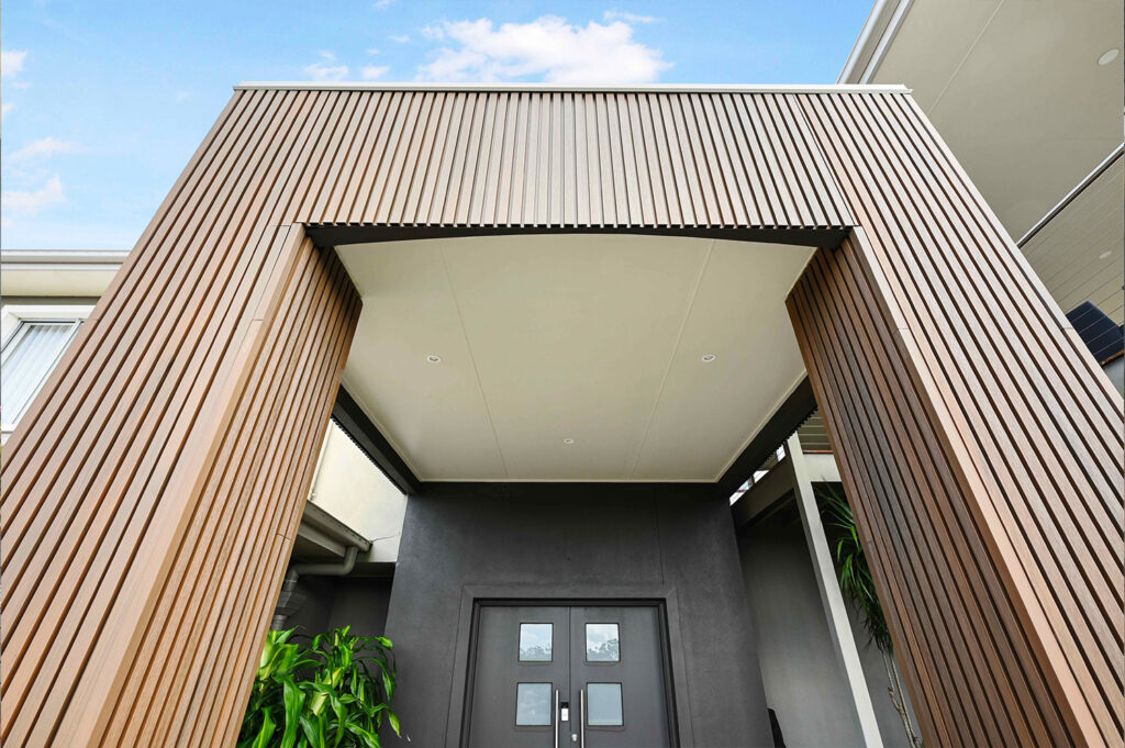 Upward angle of modern entry framed with Country Teak composite cladding, showing vertical battens and recessed lighting above dark double doors.