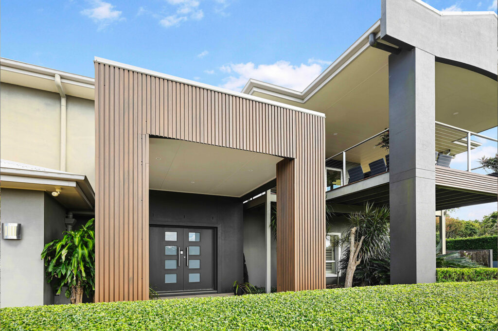 Frontal view of entrance with Country Teak composite cladding, featuring vertical battens, double doors, and surrounding hedges beneath a modern balcony.
