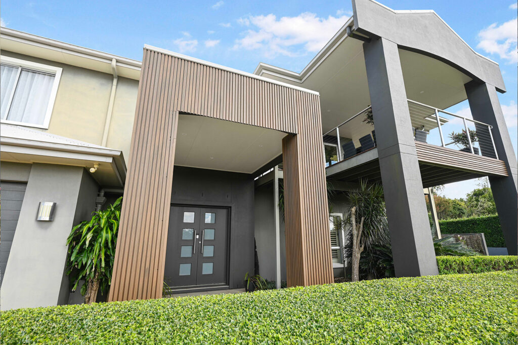 Angled view of home entry with Country Teak composite cladding, double doors, and a modern balcony framed by grey columns and lush hedges.