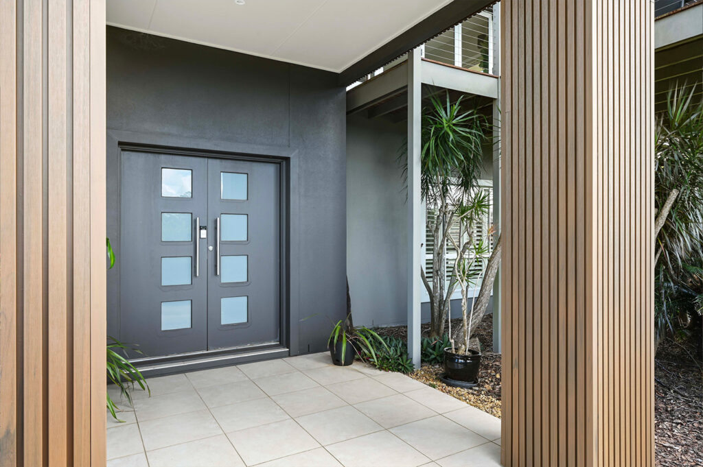 Close-up of tiled front porch with dark double doors, framed by Country Teak composite cladding and tropical potted plants.