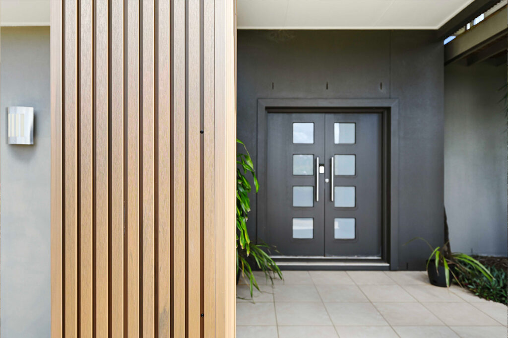 Front door entry framed by Country Teak composite cladding, showing vertical batten texture alongside tiled flooring and matte black double doors.