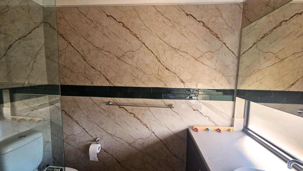 Bathroom with beige marble-patterned SPC wall panels, dark green horizontal accent strip, chrome towel rail, white toilet, frosted horizontal window, and countertop with small colorful items, creating a bright and modern space.