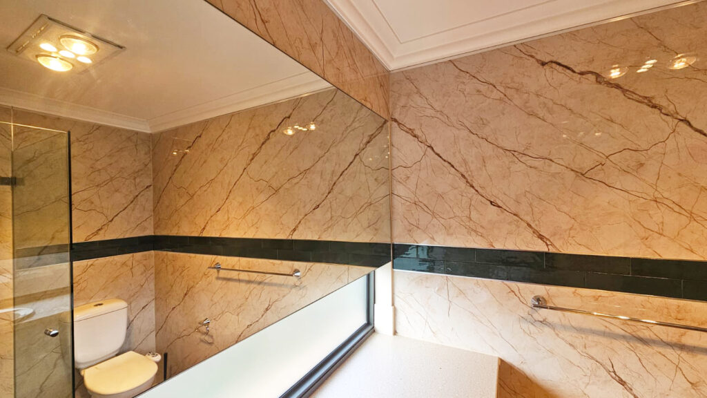 Modern bathroom with beige marble-patterned SPC wall panels, a dark green horizontal accent strip, large wall mirror, glass shower enclosure, and white toilet, illuminated by ceiling lights.