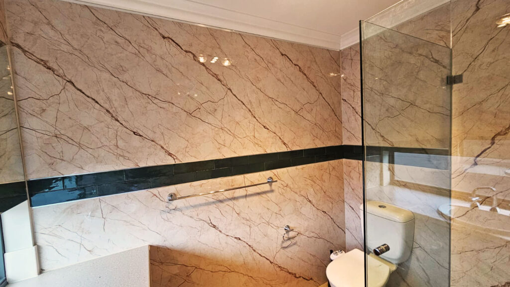 Bathroom interior with beige marble-patterned SPC wall panels, a dark green horizontal accent strip, glass shower partition, white toilet, and chrome towel rail, creating a modern and clean design.