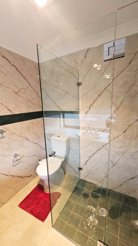 Bathroom with beige marble-patterned SPC wall panels, a dark green horizontal accent strip, glass shower enclosure, white toilet, red floor mat, and green-tiled shower floor, creating a modern and clean look.