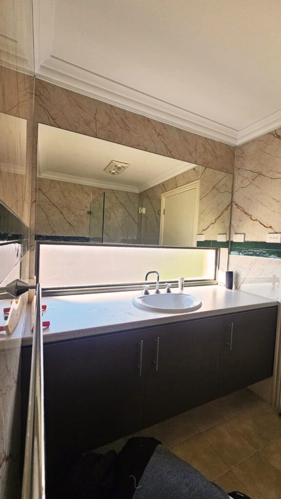 Modern bathroom vanity with a white countertop, round basin sink, chrome faucet, large mirror, and beige marble-patterned SPC wall panels with a dark green accent strip, illuminated by natural light from a frosted horizontal window.