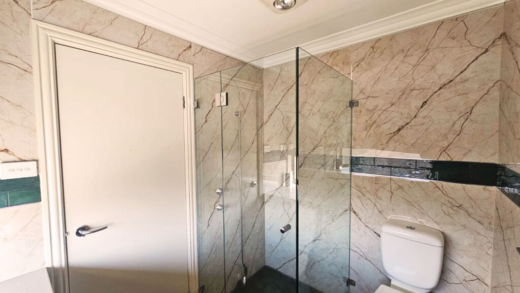 Bathroom with beige marble-patterned SPC wall panels, dark green horizontal accent strip, glass shower enclosure with chrome fixtures, white toilet, and white door, creating a sleek and contemporary design