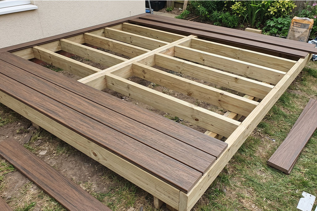 Deck Cost Artboard 6 Composite decking boards being installed over a timber frame base during deck construction in a backyard. The exposed joists and partially laid boards show the step by step process of building a durable composite decking structure.