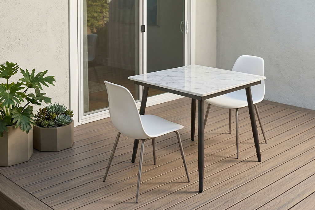 Small outdoor patio setup with a square marble top table and two white chairs placed on composite decking. Neutral walls and sliding glass doors frame the space while potted green plants add a touch of color beside the seating area.