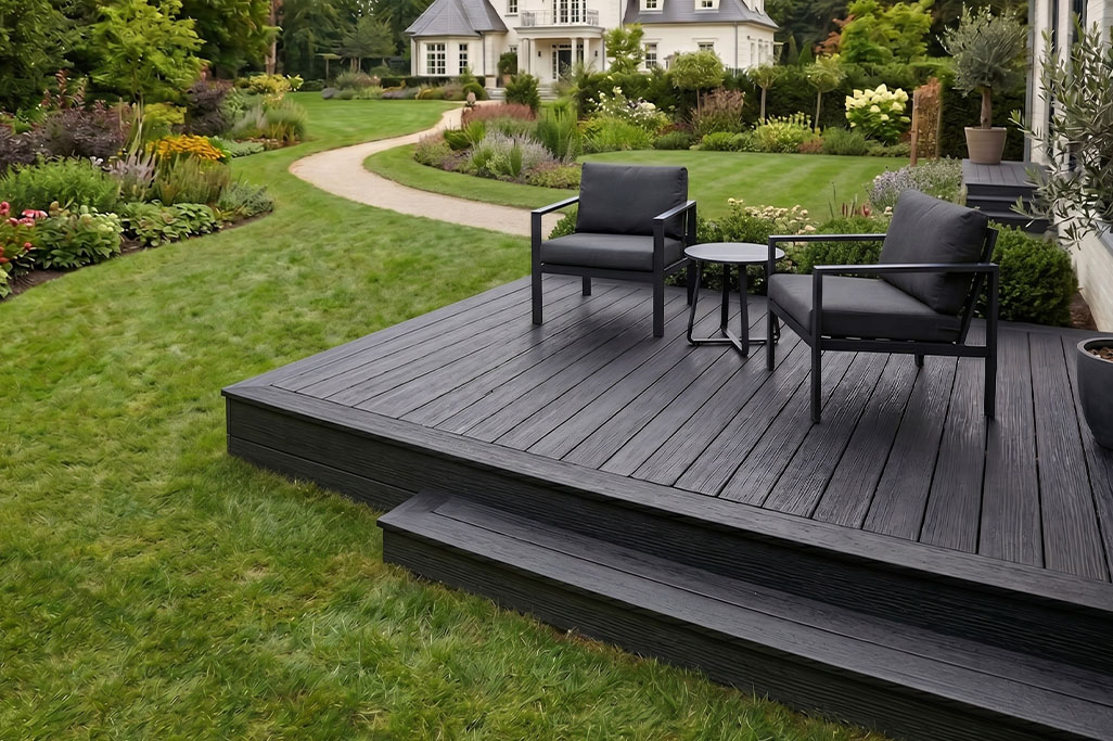 Dark composite decking patio with two cushioned chairs and a small round table overlooking a landscaped garden with a winding path and lush greenery. The raised deck creates a defined outdoor seating area that blends into the surrounding yard and elegant home.
